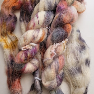 May include: Close-up of multi-coloured yarn in shades of orange, purple, cream, and grey. The yarn is loosely spun, with a soft, textured appearance. The colours blend together, creating a variegated effect, suitable for knitting or crochet projects.