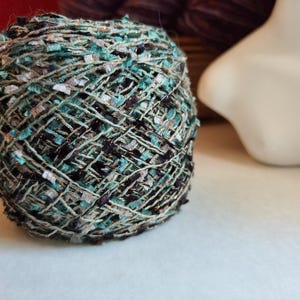 May include: A ball of textured yarn featuring a blend of teal, black, and metallic silver fibers. The yarn has a multi-strand construction with varying thicknesses and a variegated appearance, creating a complex, interwoven pattern throughout the ball.