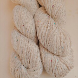 Pu&ograve; includere: Primo piano di un gomitolo di filato beige con paillettes colorate. Il filato &egrave; attorcigliato strettamente, mostrando la consistenza delle fibre. Le paillettes sono piccole e sparse nel filato, aggiungendo un tocco di brillantezza. Ideale per progetti di maglia e uncinetto.