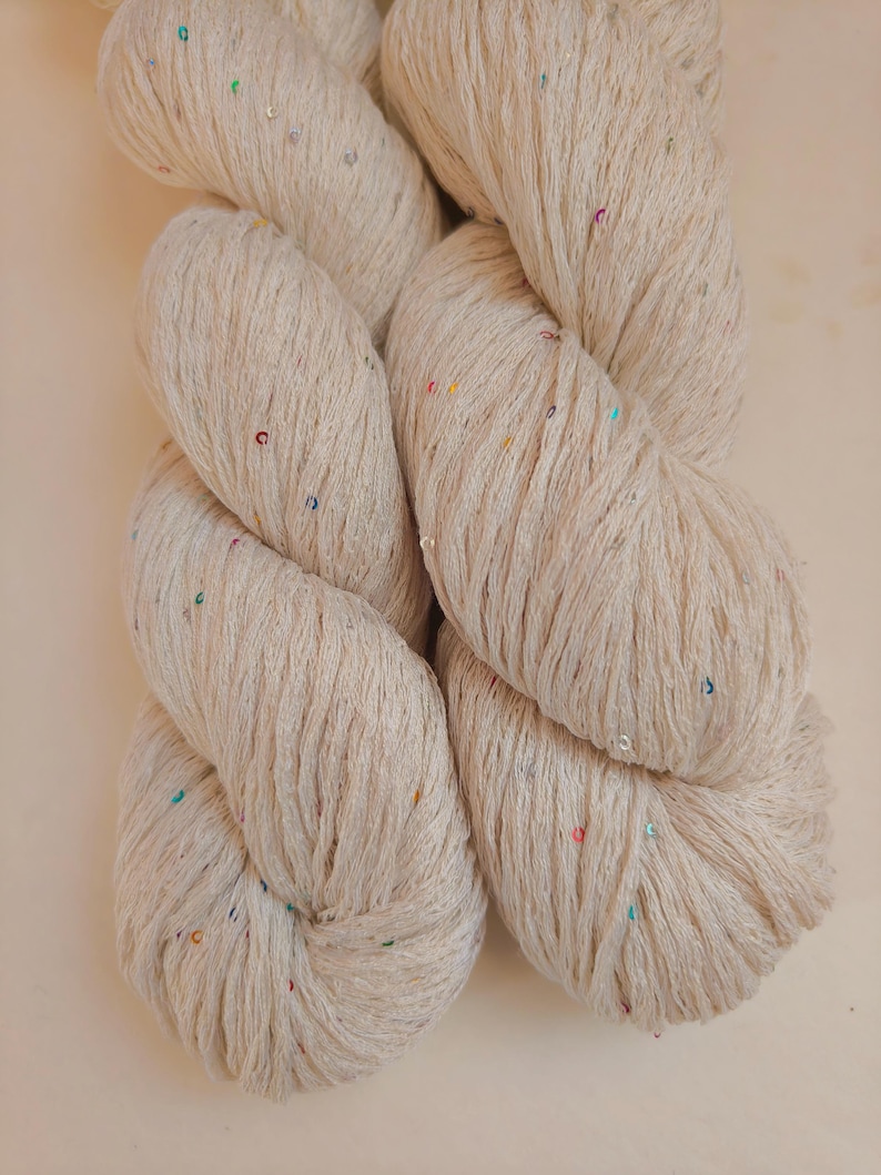 Pu&ograve; includere: Primo piano di un gomitolo di filato beige con paillettes colorate. Il filato &egrave; attorcigliato strettamente, mostrando la consistenza delle fibre. Le paillettes sono piccole e sparse nel filato, aggiungendo un tocco di brillantezza. Ideale per progetti di maglia e uncinetto.