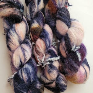 May include: Close-up of several skeins of hand-dyed yarn. The yarn is a mix of colors, including deep blues, purples, and soft pinks, with a fluffy texture. The yarn is likely used for knitting or crocheting projects.