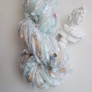 May include: A skein of light blue yarn with silver and gold metallic threads and sequins. The yarn is fluffy and textured, with a mix of colors and materials. A white bust of a classical figure is in the background.