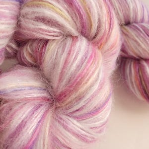 May include: Close-up of a skein of soft, colorful yarn. The yarn is a blend of pink, white, and purple hues, with subtle hints of yellow and a shimmering, iridescent quality. The texture appears fluffy and delicate, ideal for knitting or crocheting projects.