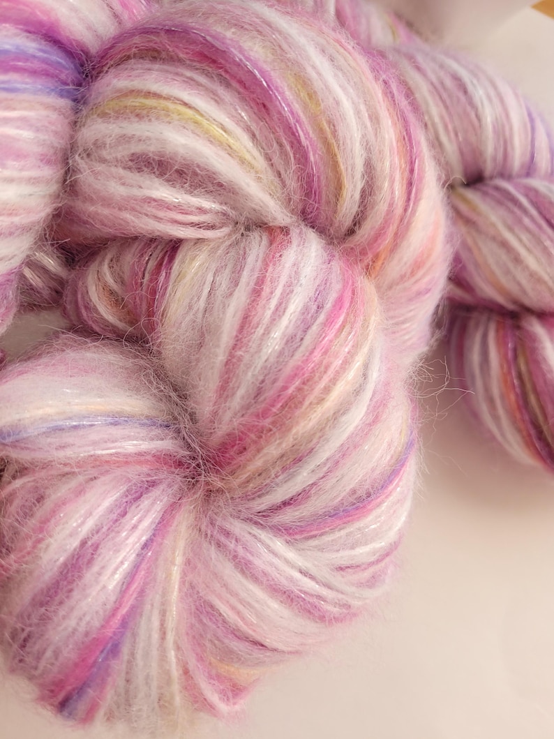 May include: Close-up of a skein of soft, colorful yarn. The yarn is a blend of pink, white, and purple hues, with subtle hints of yellow and a shimmering, iridescent quality. The texture appears fluffy and delicate, ideal for knitting or crocheting projects.