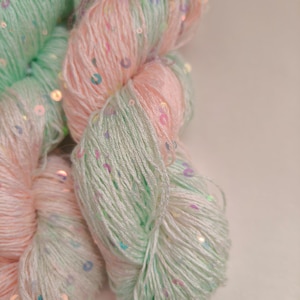 Peut inclure: Gros plan sur une pelote de laine aux couleurs pastel. Le fil est un mélange de rose clair et de vert menthe, parsemé de paillettes irisées. La laine semble douce et duveteuse, adaptée aux projets de tricot ou de crochet.
