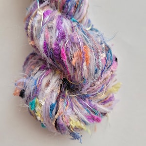 May include: A close-up of a colorful yarn ball. The yarn is a mix of purple, pink, blue, and yellow threads with a textured, multi-layered appearance. The yarn appears to be a blend of different materials, including some with a shiny, metallic finish.