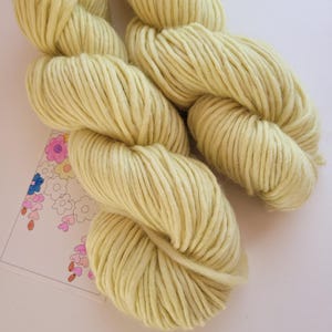 Peut inclure: Gros plan sur deux écheveaux de fil jaune pâle. La laine épaisse est étroitement enroulée, montrant des brins individuels. Le fil repose sur une surface blanche avec un motif floral en arrière-plan. Idéal pour les projets de tricot et de crochet.