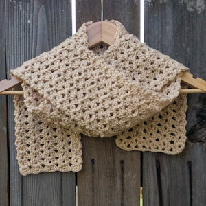 Beige scarf in a shell pattern on a hanger in front of a dark wooden fence.