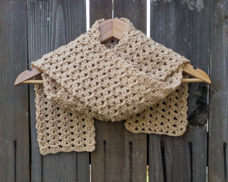 Beige scarf in a shell pattern on a hanger in front of a dark wooden fence.