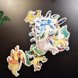 May include: A collection of colorful stickers featuring various cartoon characters. The stickers showcase a variety of creatures in shades of blue, yellow, green, orange, and pink. The stickers are on a black surface.