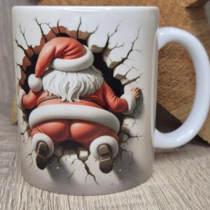 Könnte beinhalten: Weiße Keramik-Tasse mit einer 3D-Illustration von Santa Claus, der aus einer Wand bricht. Santa trägt einen roten Anzug mit weißer Verzierung und passendem Hut. Die Tasse hat einen weißen Henkel und ein weihnachtliches Thema.