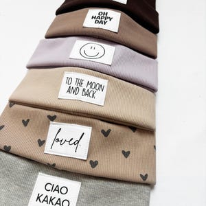 Könnte beinhalten: Ein Stapel aus sechs gerippten Mützen in verschiedenen Farben wie Dunkelbraun, Hellbraun, Helllila, Beige und Grau. Jede Mütze hat ein weißes rechteckiges Etikett mit schwarzem Text oder Grafiken, darunter Schriftzüge wie mini BOSS, OH HAPPY DAY, TO THE MOON AND BACK, loved und CIAO KAKAO sowie ein Smiley-Gesicht und ein Herzmuster.