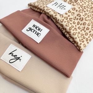 Könnte beinhalten: Drei übereinander liegende Mützen in Beige- und Rosatönen. Eine ist mit einem Leopardenmuster versehen. Jede Mütze hat ein weißes Etikett mit schwarzem Text. Die Etiketten lauten "Milla", "moin giorno" und "hej."