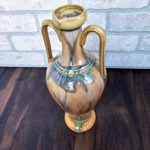 May include: A brown and green ceramic vase with two handles and a decorative band around the middle. The vase has a textured, glazed finish.