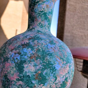 May include: A decorative, hand-painted vase with a bulbous body and a slender neck. The vase is primarily teal green, with splashes of pink, blue, and bronze-colored paint. The textured surface gives it a unique, artistic appearance.