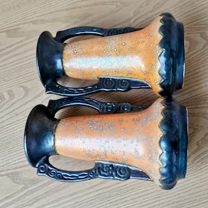 May include: Two orange ceramic vases with black handles and decorative details. The vases have a speckled glaze and a scalloped rim.