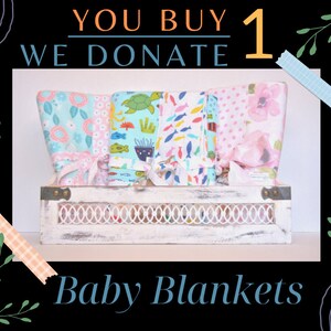 May include: A wooden crate filled with five colorful baby blankets. The blankets feature different patterns, including flowers, fish, and sea turtles. The text "You Buy 1 We Donate 1" is displayed above the crate.  Baby Blankets.