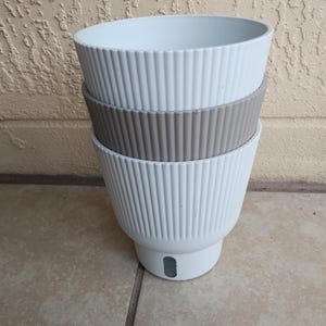 May include: Three stacked, ribbed, ceramic planters. The top and bottom planters are white, and the middle one is a muted gray. Each planter has a wide, open top and a drainage hole near the base. The planters are on a tiled surface.