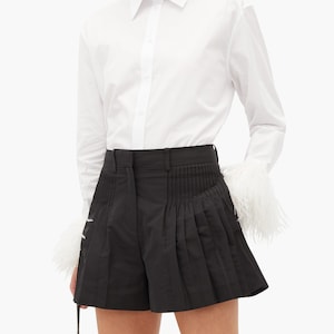 May include: A white long-sleeve button-down shirt with feather trim on the cuffs paired with black pleated shorts.