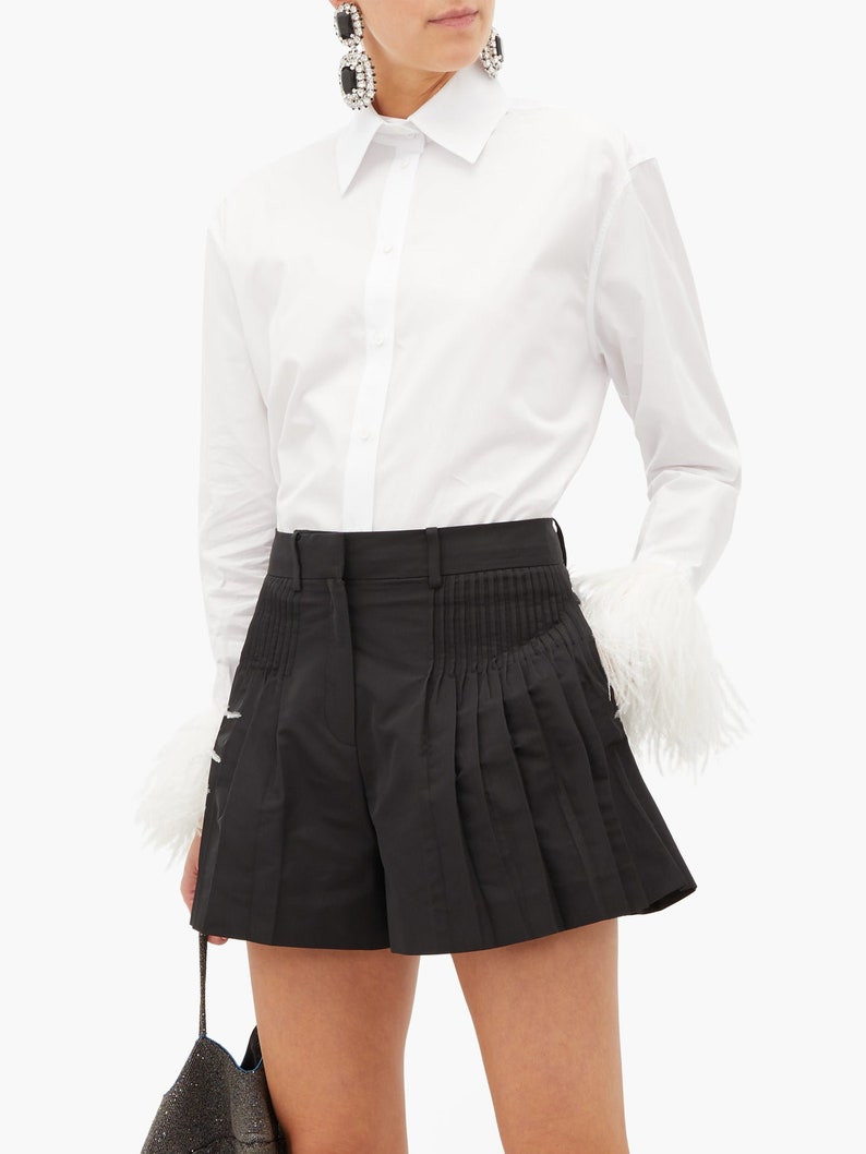May include: A white long-sleeve button-down shirt with feather trim on the cuffs paired with black pleated shorts.