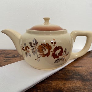 Peut inclure: Une théière de couleur crème avec un motif floral brun. La théière a un couvercle et une poignée.