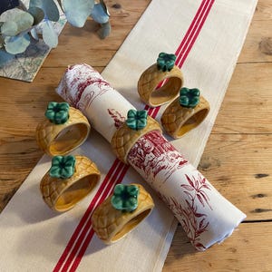May include: Six pineapple-shaped napkin rings with green leaves on a white linen tablecloth with red stripes. The napkin rings are made of ceramic and are a light brown color.