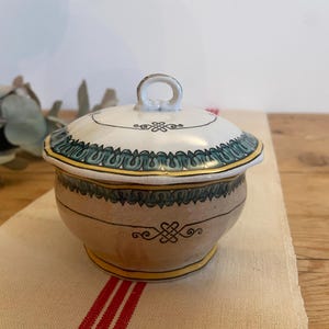 May include: A small, round, ceramic jar with a lid. The jar is decorated with green and yellow floral patterns and a brown and white geometric design. The lid has a small knob on top.