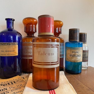 May include: A selection of vintage apothecary bottles in shades of blue and amber. The bottles feature labels with French text and are topped with cork or metal closures. They are displayed on a wooden surface.