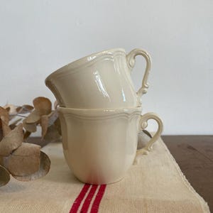 Peut inclure: Deux tasses en céramique couleur crème empilées avec une finition brillante, présentant un détail de bord festonné et une anse incurvée. Les tasses ont un design simple avec une surface lisse, affichant un ton beige clair uniforme.