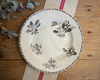 May include: A large, white ceramic plate with a brown floral design. The plate has a raised rim and a scalloped edge. The flowers are stylized and have a vintage look.