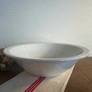 Può includere: Una ciotola in ceramica bianca con un bordo largo, appoggiata su un panno a righe bianche e rosse. La ciotola ha una finitura liscia e lucida e un design semplice e classico. La ciotola viene probabilmente utilizzata per servire o preparare il cibo.