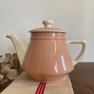 Op de afbeelding: Een keramische theepot met een glanzende pastelroze behuizing en een crèmekleurige tuit, handvat en deksel. Het deksel heeft een kleine, ronde knop aan de bovenkant. De theepot heeft een brede, taps toelopende voet en een gladde, reflecterende afwerking.