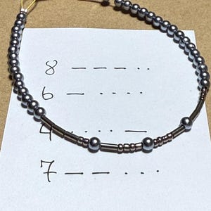 May include: A beaded bracelet with gray beads and gold-colored clasp. The bracelet is on a white paper with handwritten numbers and Morse code symbols. The bracelet is a delicate piece of jewelry.