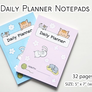 May include: Two daily planner notepads with cute cat illustrations. The notepad on the left is light blue with a white cat and a yellow yarn ball. The notepad on the right is light purple with a white cat and a gray cat. Both notepads have the text "Daily Planner" and "MONTH:" printed on them. The notepads are 5 inches wide by 7 inches tall and have 32 pages.