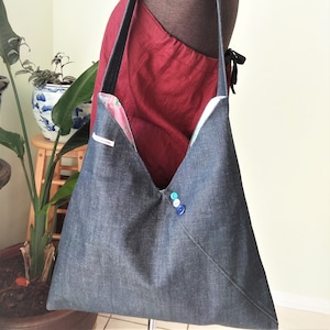 Peut inclure: Un sac cabas en denim bleu foncé avec une longue bandoulière. Le sac a un rabat avec trois boutons dans des tons de bleu et de vert.