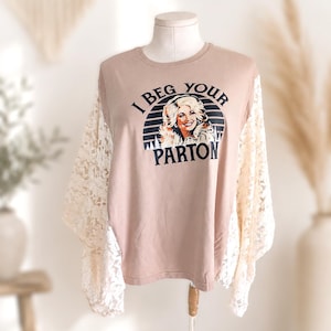 May include: A tan long-sleeve shirt featuring a graphic print of a woman with blonde hair and the text I BEG YOUR PARTON centred on the chest. The shirt has long, loose-fitting white lace sleeves with a floral pattern and a crew neckline.