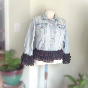 May include: A light blue denim jacket featuring a classic collar, button-front closure, and two chest pockets. The hem and sleeve cuffs are accented with layers of black ruffled tulle fabric, creating a distinct contrast against the light wash denim.
