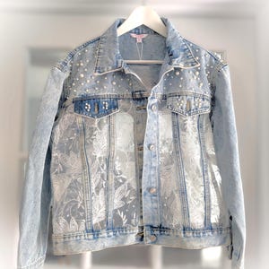 Peut inclure: Une veste en jean bleu clair délavé à l'acide avec des panneaux en dentelle florale blanche sur le devant, un col classique, des manches longues et des boutons. Le haut de la poitrine et les poches sont ornés de perles blanches.