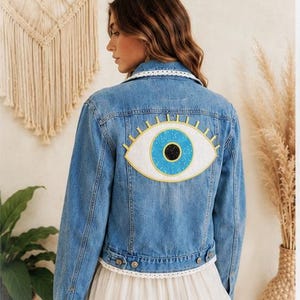 May include: A light blue denim jacket with a large embroidered eye design on the back. The eye features a blue iris, a white background, and gold-colored eyelashes. The jacket has a white lace trim on the collar and hem.