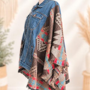 May include: A blue denim jacket featuring a brown, red, and turquoise patterned textile back and side panels. The garment has a classic collar, button-front closure, chest pockets, and a fringed hemline along the lower edge of the patterned fabric.