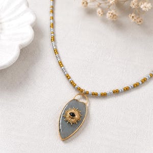 May include: A necklace featuring a teardrop-shaped grey stone pendant with a gold-toned sunburst emblem and a central black stone. The chain consists of alternating small metallic gold and silver beads arranged in a repeating pattern along the strand.