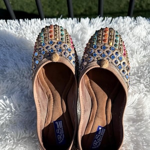 May include: A pair of beige flat shoes with colorful beaded embellishments. The shoes have a rounded toe and a small, round button on the top. The soles are labeled "R&S" and "Jutti Stone".