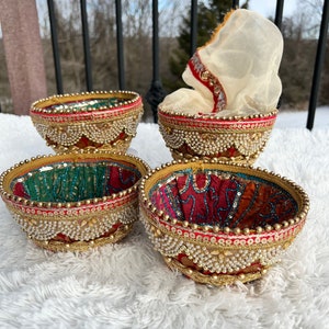 May include: Four decorative bowls with intricate designs. The bowls are made of a red, gold, and green fabric with pearl accents. The bowls are arranged on a white fluffy surface.