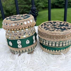 May include: Two round green and gold decorative boxes with intricate embroidery and embellishments. The boxes are made of fabric and have a traditional Indian design.