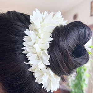 May include: A white flower hair accessory made of fabric petals, worn in a bun of dark hair.