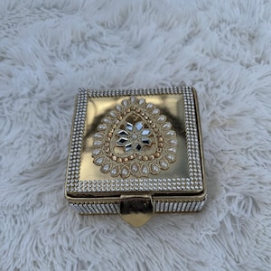Könnte beinhalten: Eine goldene quadratische Box mit einem dekorativen Design in Form eines Tränen tropfens mit weißen Perlen und einer verspiegelten Mitte. Die Box ist mit einem silbernen Perlenrand verziert.