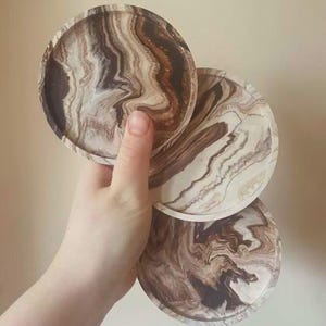 May include: Three circular coasters featuring a swirling marbled pattern in shades of dark brown, cream, and tan. Each coaster has a raised edge and a smooth, glossy finish, displaying a unique fluid design that mimics natural stone textures.