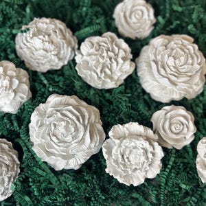 May include: A collection of white, sculpted floral decorations, resembling roses and peonies, are arranged on a bed of green, shredded paper. The flowers vary in size and detail, showcasing intricate petal designs.