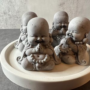 May include: Four gray laughing Buddha figurines with rounded faces and hands clasped in prayer. The figurines are sitting on a white circular tray.
