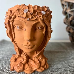 Puede incluir: Maceta de terracota con forma de cabeza de mujer con una corona floral y detalles de piedras preciosas blancas. La maceta tiene un rostro y un cabello detallados, y la base está decorada con hojas.
